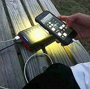 a solar charger laying on a table with a cable connecting it to a device; the device is being held so that the light on the back is shining on the solar panel to recharge it.
