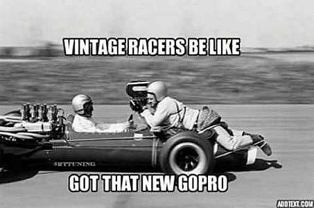 a vintage race car being driven at high speed; a man with a large video camera is balanced on the hood, aiming the camera on the driver's face. Caption: Vintage racers be like, "Got that new GoPro!"