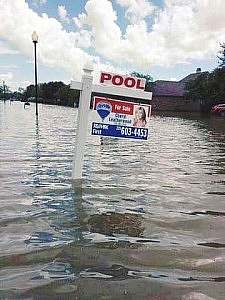 In the middle of a flooded neighborhood where only the tops of houses and streetlights rise above the water, pokes up a real estate sign offering for sale a house with a pool