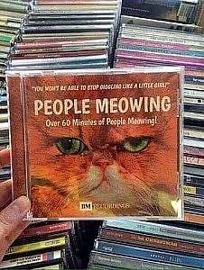 Stacks of CDs in the background. Someone holding a CD in front with the face of an orange cat on the cover called, "People Meowing" Over 60 Minutes of People Meowing! "You won't be able to stop giggling like a little girl!