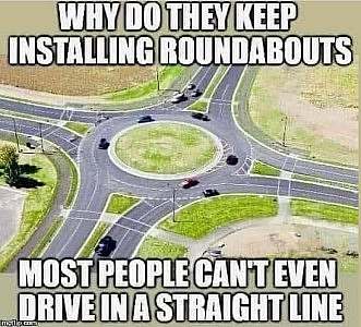An aerial view of a traffic roundabout with four lanes entering and exiting at 90 degrees. Caption: Why do they keep installing roundabouts? Most people can't even drive in a straight line!