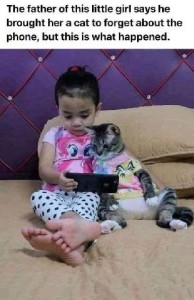 An adorable little girl - maybe 3 years old - is sitting on a bed looking at something on a smartphone. Next to her is a cat, also leaning back against pillows, wearing an little outfit, also watching the screen. Caption: The father of this little girl says he brought her a cat to forget about the phone, but this is what happened.