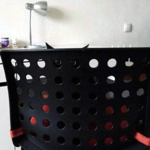 A photo of a black laundry basket with a black cat inside, his green eyes peering through two of the holes and the tips of his ears showing above the top.