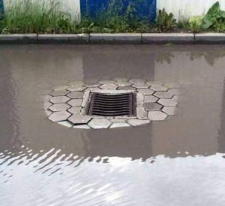 A photo of a flooded street. In the center is the drain cover which had been installed with a higher ring of the street around it; it is high and dry.