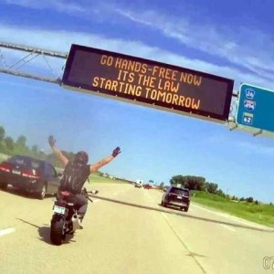 Photo, taken from behind, of a motorcyclist on a freeway with both hands up in the air. Above him is a digital roadsign that says, "Go hands-free now. It's the law starting tomorrow."