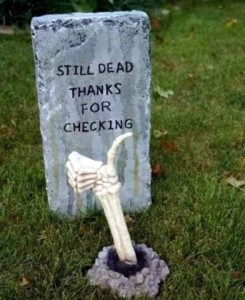 Photo of a headstone with the words, "Still dead, thanks for checking" and a skeletal hand coming up through the ground in front of it with the thumb up