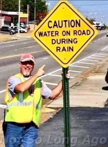 A construction worker pointing to a yellow sign on a road that says, "Caution water on road during rain."