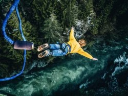 A man high over a rapidly moving river, attached to bungee cords, facing downward with arms outstretched.