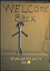 A piece of cardboard with the hand-printed words "Welcome back" at the top. Below that is a stick-man made of cotter pins. Caption: If you get this you're old
