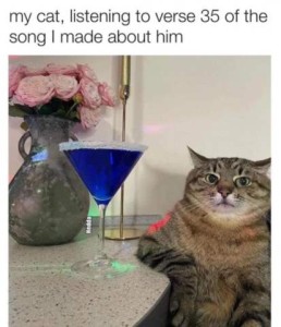 A grey tabby cat sitting at a counter, sat up "human style", his right elbow on the counter, looking bored. There is a martini glass with a blue drink next to his elboe, and a vase of flowers in the background. Caption: My cat, listening to verse 35 of the song I made about him.