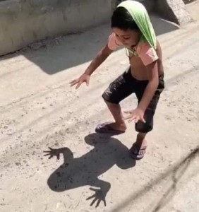 photo of a small child, in the sun, his shirt pulled up over the back of his head, arms and fingers outstretched; in front of him is a shadow of a frog