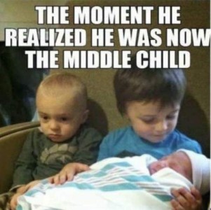 A preschool-aged boy holding and smiling down at a newborn baby. To his left sits his little brother, looking glum. Caption: The moment he realized he was now the middle child.