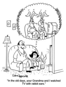 Cartoon of an older man sitting on a davenport with a little girl, a television set in front of them. He is telling her, "In the old days, you Grandma and I watched TV with rabbit ears." In a thought bubble over the little girl's head is the same old man, with an old woman, sitting in front of the television set wearing hats with big rabbit ears on them.
