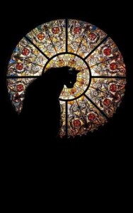 Striking photo of an elaborate circular stained-glass window surrounded in black, with a black cat silhouetted in front of it.