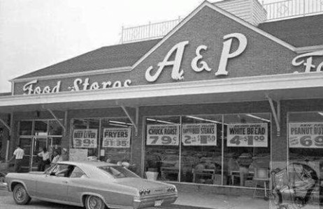 A photo of an A&P with some signs in the window including white bread 4 for $1.00, peanut butter for 69¢, chuck roast for 79¢ a pound, chicken fryers 31 to 35¢.