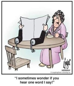 Cartoon of a woman in a robe and curlers sitting at the breakfast table. Across from her is a newspaper being held up ,as if being read, by two hands on fake arms clamped to the table behind it. Caption: "I sometimes wonder if you hear one word I say!"