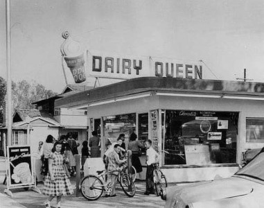 A vintage black and white photo of a Dairy Queen with kids standing around the entrance, and on bikes.
