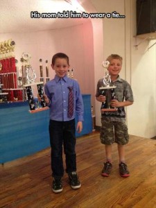 Two young boys holding trophies, standing in what looks to be school gym. One is wearing camo shorts and shirt. The other is wearing long dark trousers and a blue dress shirt with a necktie hanging from the pocket. Caption: His mom told him to wear a tie...