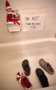 Photo of a bathtub with multiple croc shoes facing the built-in soap holder. An Elf is sitting on the soap holder holding a little bag of Goldfish crackers. Next to him on the wall is a sign saying, "Don't feed the crocs."