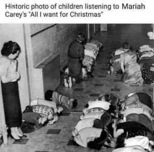 50's or 60's photo of children lined up in the hallway of a school, hunched down on the floor with their hands clasped behind their necks while teachers stand by and watch them participate in the tornado drill. Caption: Historic photo of children listening to Mariah Carey's "All I Want For Christmas"