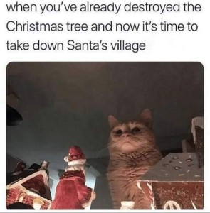 Yellow cat sitting on a high shelf, near the ceiling, surrounded by Christmas decorations; his face is lit from the bottom, giving him a sinister look. Caption: When you're already destryed the Christmas tree and now it's time to take down Santa's village.