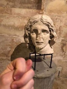 Photo of a bust of a woman; the nose is broken off. In front of her someone is holding his hand in a fist with his thumb between his index and middle fingers.