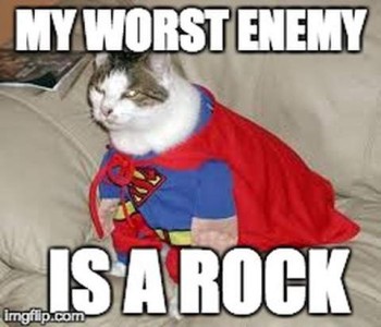 White and gray cat sitting on a beige upholstered furniture piece, wearing a Superman costume with a red cape. Caption: My worst enemy is a rock.
