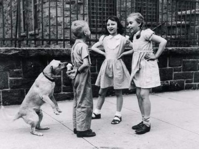 Old black and white photo of a little boy standing on the sidewalk talking to 2 little girls while he hides an ice cream cone behind his back. Unbeknownst to him, apparently, a dog is eating his ice cream.