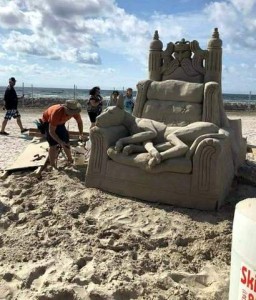 Photo of a sunny day at the beach where a man is working on an elaborate sand sculpture of a chair with what looks to be a throne back behind it, with a royal person in the middle and pillars on the sides. There is a dog laying on the chair, head on the arm. May be a great dane.