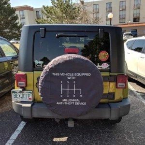 Photo of the back of a yellow Jeep with a black hardtop. The cover over the spare tire shows a diagram of the 6 speeds of a manual transmission, and the words, "This vehicle equipped with millennial anti-theft device.