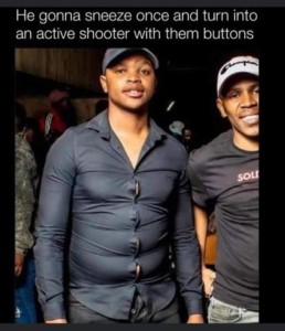Photo of 2 young men standing in the photo, both are wearing ball caps, one is wearing a t-shirt and the other is wearing a button-down shirt that is clearly too small and has large gaps between each button; both of them are smiling. Caption: He gonna sneeze once and turn into an active shooter with them buttons!