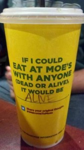 Photo of a yellow cardboard drink cup sitting on a table. On the side is printed "If I could eat at Moe's with anyone (dead or alive) it would be..." and a line where someone could write in an answer. Someone had written in, "ALIVE"