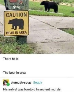 Photo of a black bear on a lawn in the background; a sign on a pole in the foreground that says, "Caution bear in area" with a picture of a black bear; they are both standing and facing the left. Caption: There he is. The bear in area. Comment: His arrival was foretold in ancient murals.